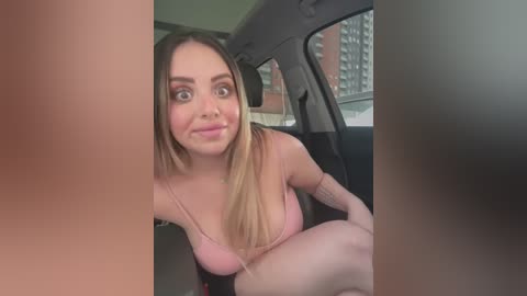 Media: Video of a young, fair-skinned woman with long, straight blonde hair, wearing a pink spaghetti-strap top, sitting in the backseat of a car. The background shows a cityscape with high-rise buildings.