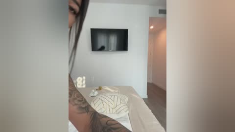 A video of a woman with tattoos, partially visible, in a modern bedroom with white walls, a flat-screen TV, and a bed with a cream-colored sheet and a geometric-patterned pillow.