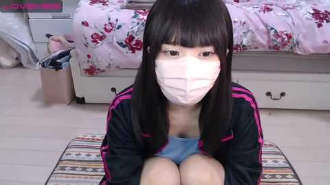 Media: A video of an Asian woman with long black hair, wearing a pink surgical mask, blue tank top, and black jacket, sitting on a striped rug in a cluttered bedroom with a pink floral bedspread.