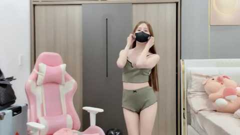 Media: Video of a slender, fair-skinned woman with long brown hair wearing a green crop top and matching shorts, black face mask, and pink gaming chair in a modern bedroom with a beige bed and wooden wardrobe.