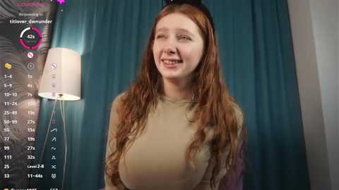 Media: A video of a smiling young woman with long, wavy red hair, wearing a beige sweater, standing against teal curtains.