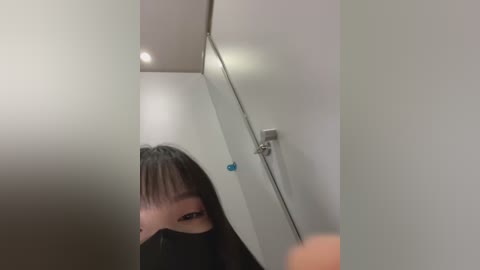 Media: Video of a young Asian woman with long, dark hair, wearing a black face mask, standing in a narrow, white-walled corridor with a reflective mirror and a blue object hanging on the wall.