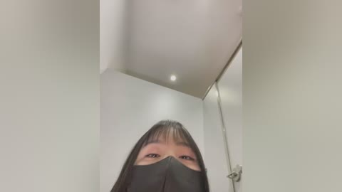 Media: A video of an Asian woman with long black hair and bangs, wearing a black face mask, taken from a low angle, revealing her eyes and nose, in a modern, white-walled room with recessed lighting and a metal rail.