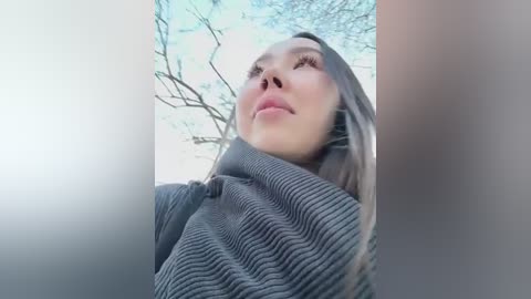 Media: Video of a young Asian woman with long, straight hair, wearing a ribbed gray sweater, looking upward towards the sky with bare tree branches in the background.