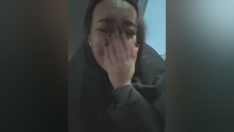 Media: A video shows a young woman with pale skin, dark hair, and dark eyes, covering her face with her hand, wearing a black jacket. She is seated in a dimly lit room, with a blurry background.