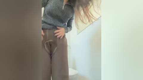 Media: Video of a woman with long, wavy hair, wearing a dark gray, ribbed sweater and high-waisted, gray pants, standing in a dimly lit room with a white wall and a beige door.