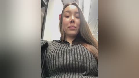 Media: Video of a young woman with long, straight, light brown hair, wearing a gray, ribbed sweater, seated in a dimly lit airplane cabin. Her expression is neutral, and her body is slightly angled towards the camera.