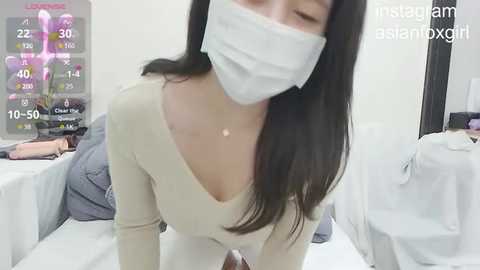 Media: A video of a woman with long black hair, wearing a white mask, leaning forward, with a text overlay. She has a slim figure and is wearing a beige long-sleeve top. The background features a medical setting with white beds.