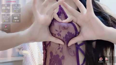 Media: Video of a woman with fair skin, long dark hair, wearing a sheer purple lace bra. Her hands form a heart shape over her breasts, revealing the bra's intricate design.