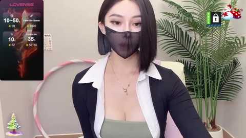 Media: Video of an East Asian woman with a bob haircut, wearing a black mask, green top, black cardigan, and a white blouse, sitting in a modern office with a fern plant.