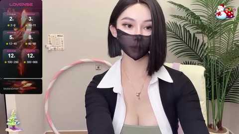 Media: A video of a young East Asian woman with a bob haircut, wearing a black mask, black blazer, and a low-cut white shirt, in an indoor setting with a plant and a calendar.