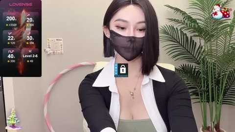 Media: Video of an East Asian woman with straight black hair, wearing a black face mask, black blazer, and white shirt. Background includes a plant and a clock.