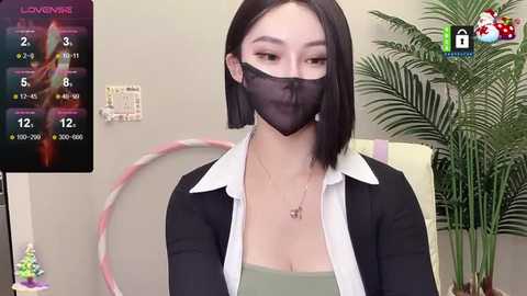 Media: Video of an East Asian woman with short black hair, wearing a black face mask, white blouse, and black cardigan, seated in front of a green plant.