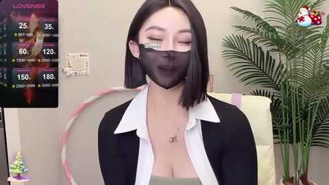 Media: A video of an Asian woman with straight black hair, wearing a black mask, white blouse, and black cardigan, standing indoors near a plant, with a TV displaying a live stream.