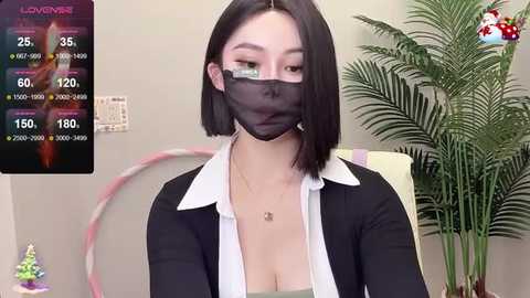 Media: Video of a young East Asian woman with straight black hair, wearing a black face mask, white shirt, and black cardigan, seated indoors with a potted plant behind her.