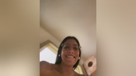 Media: A low-angle video of a smiling, topless Asian woman with long black hair and glasses, standing in a bathroom with beige walls and a toilet roll holder.