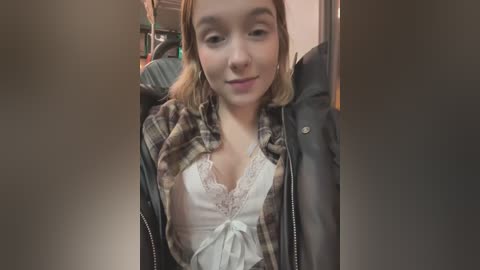 Media: Video of a young woman with light skin, shoulder-length blonde hair, and a slight smile, wearing a white lace top under a plaid shirt, sitting in a car.