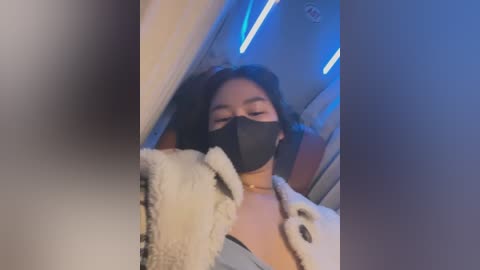 Video of a young woman with long, dark hair, wearing a black face mask and a white, fluffy robe. She appears to be indoors with blue LED lights in the background.