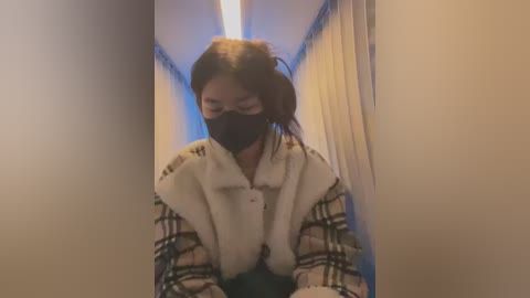 Media: A video of an Asian woman with long black hair, wearing a black face mask, a white fur-trimmed jacket, and a plaid shirt, standing in a narrow hallway with beige walls and a blue light.