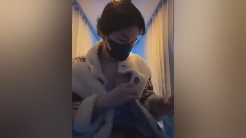 Media: Video of an East Asian woman in a cozy hallway with dim lighting, wearing a white robe, black mask, and hair tied up, looking at a smartphone, partially blurred background.