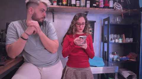 Video of a bearded man in a gray shirt and white pants sitting pensively, and a curly-haired woman in a red sweater and patterned skirt, focused on her phone, in a cluttered, dimly-lit bar.