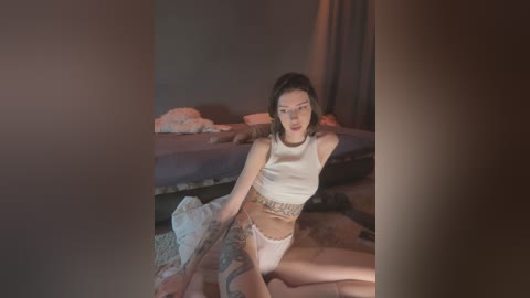 A video of an Asian woman with short black hair, wearing a white crop top and lace panties, sitting on a rug in a dimly lit bedroom with rumpled sheets and a blue bed.