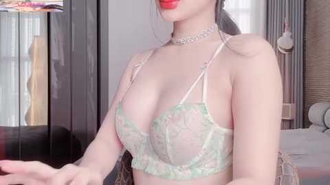 Video of an East Asian woman with fair skin, wearing a pale green lace bra revealing her large breasts and a choker necklace, standing indoors in a dimly lit room with a bed and wardrobe visible.