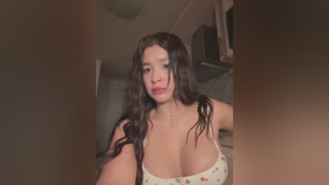 Media: A video of a young woman with long, wavy black hair and fair skin, wearing a white tank top with floral patterns. She stands in a dimly lit room with beige walls and a ceiling fan.