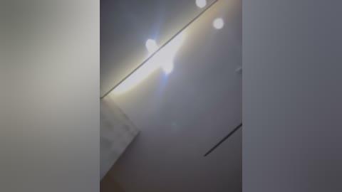 A dimly lit video showing a narrow, angular hallway with white walls, a single overhead light emitting soft white light, and a black, vertical shadow on the right wall.