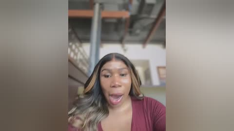 Media: Video of a young woman with medium skin tone and long, wavy hair. She wears a maroon top, and her mouth is open, suggesting surprise or excitement. The background shows an industrial-style room with exposed pipes and beams.