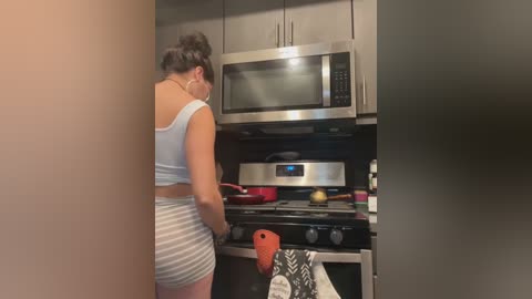 Media: Video of a woman with dark hair in a bun, wearing a white tank top and gray shorts, cooking in a modern kitchen with stainless steel appliances and a microwave.