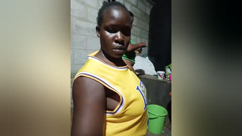 Media: Video of a muscular black woman in a yellow sports jersey, with short hair, standing indoors near a gray brick wall and green bucket.