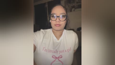 Video of a young Asian woman with glasses, wearing a white T-shirt with \"Inliterally just a girl\" and a pink ribbon, sitting indoors.