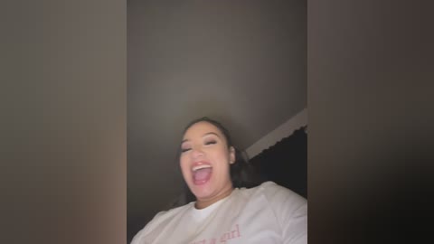 Video of a young woman with long black hair, wearing a white T-shirt, laughing joyfully. The background is dimly lit with dark, shadowy tones.