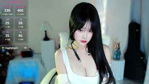 Media: Video of a young Asian woman with long black hair, wearing a white off-shoulder dress, in a modern, dimly-lit bedroom. Background includes a bed, nightstand, and digital screen displaying temperature and time.