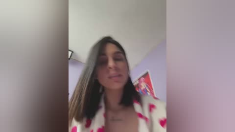 Media: Video of a Latina woman with straight, shoulder-length dark hair, wearing a white robe with pink heart patterns, standing in a room with light purple walls. She has a tattoo on her chest.