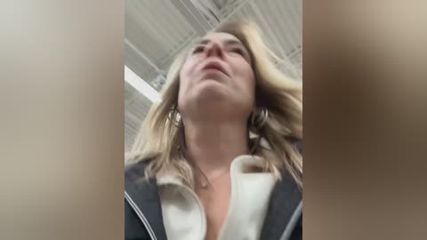 Media: A low-angle, close-up video of a blonde woman with fair skin, wearing a white shirt under a dark blazer, standing in a bright, industrial room with visible ceiling tiles.