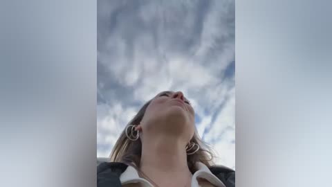 A video of a blonde woman with light skin, wearing large silver hoop earrings and a black jacket, gazing upward at a cloudy sky.