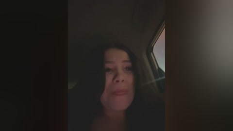 Media: A dimly lit, grainy video of a woman with long, dark hair, wearing a black top, seated in a car, looking straight ahead. The background is dark and indistinct.