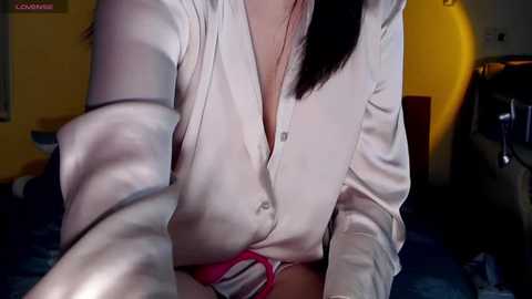 Media: Video of a light-skinned woman in a white button-down shirt, revealing cleavage, leaning forward on a dark bed. Background includes a yellow chair and dimly lit room.