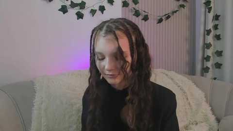 Media: Video of a young woman with long, wavy brown hair, wearing a black turtleneck, sitting on a beige couch with a fluffy white blanket, surrounded by fake ivy and purple lighting.