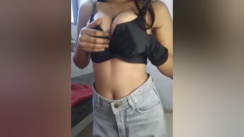 Media: Video of a woman with medium skin tone, wearing a black bra and light blue denim shorts, standing indoors near a bed with a red blanket.