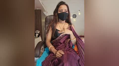 Media: Video of a South Asian woman with long dark hair, wearing a black face mask, purple sari, and gold jewelry, seated on a brown leather chair in a cozy, well-lit room.