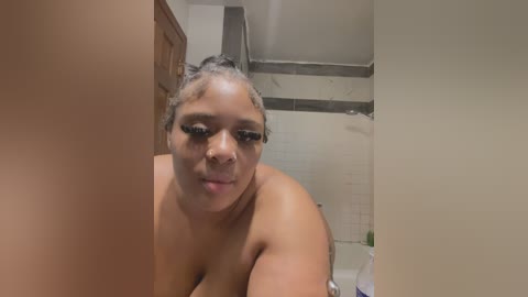 Media: A video of a plus-sized, light-skinned Black woman with braids, wearing heavy makeup and nude in a bathroom with tiled walls, looking directly at the camera.