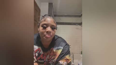 Media: Video of a Black woman with braided hair, wearing a tie-dye shirt, leaning over a sink in a small, dimly lit bathroom with white walls and a tiled shower.