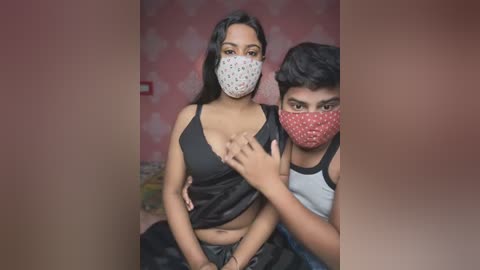 Media: Video of a young South Asian woman and man in masks, the woman wearing a black camisole, the man a tank top, both sitting on a bed.