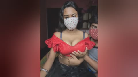 Media: Video of a woman with medium brown skin, wearing a red off-the-shoulder top, revealing cleavage, and a polka-dot mask, surrounded by two masked men in a dimly lit room.