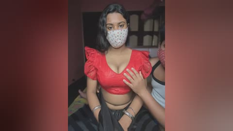 Media: Video of a South Asian woman with medium skin tone, black hair, wearing a red polka-dotted mask, red lace blouse, and black skirt, in a dimly-lit room with shelves in the background.