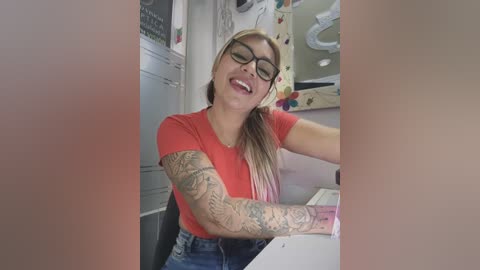 Media: Video of a smiling, tattooed, blonde woman with glasses, wearing a red T-shirt and blue jeans, sitting at a table in a brightly lit room.