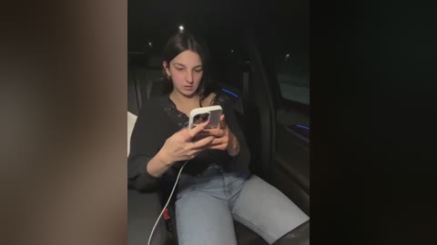 Media: A video shows a young woman with long black hair, wearing a black top and light jeans, seated in a car, focused on her smartphone. The car's interior is dimly lit, and outside is dark.